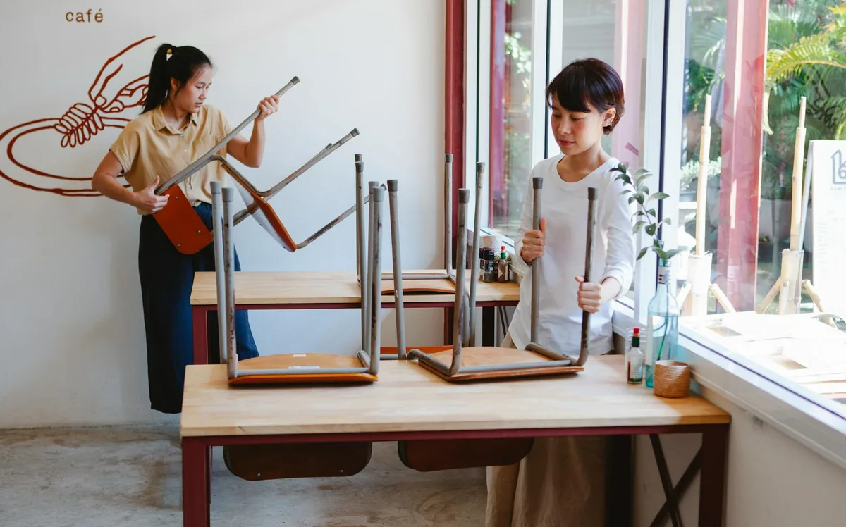 Two Asian women preparing a café for the day, arranging tables and chairs.